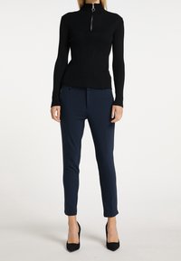 Zwart geribbeld topje met een hoge kraag en een ritsdetail aan de voorkant, gecombineerd met op maat gemaakte marineblauwe broek, afgewerkt met zwarte hoge hakken.