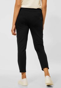 Person i svarta slim-fit byxor, vita slip-on skor och en ljusbeige topp, med ryggen vänd mot en enfärgad ljus bakgrund.