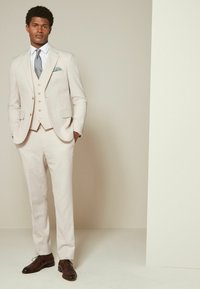 Costume beige clair à coupe cintrée, avec une veste à simple boutonnage, un gilet à trois boutons et des chaussures marron à lacets. Détail de pochette texturée.