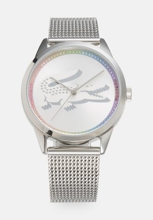 Relógio com bracelete em malha prateada e caixa metálica. O mostrador branco apresenta um bisel de cristais coloridos e um discreto logótipo de crocodilo ao centro.