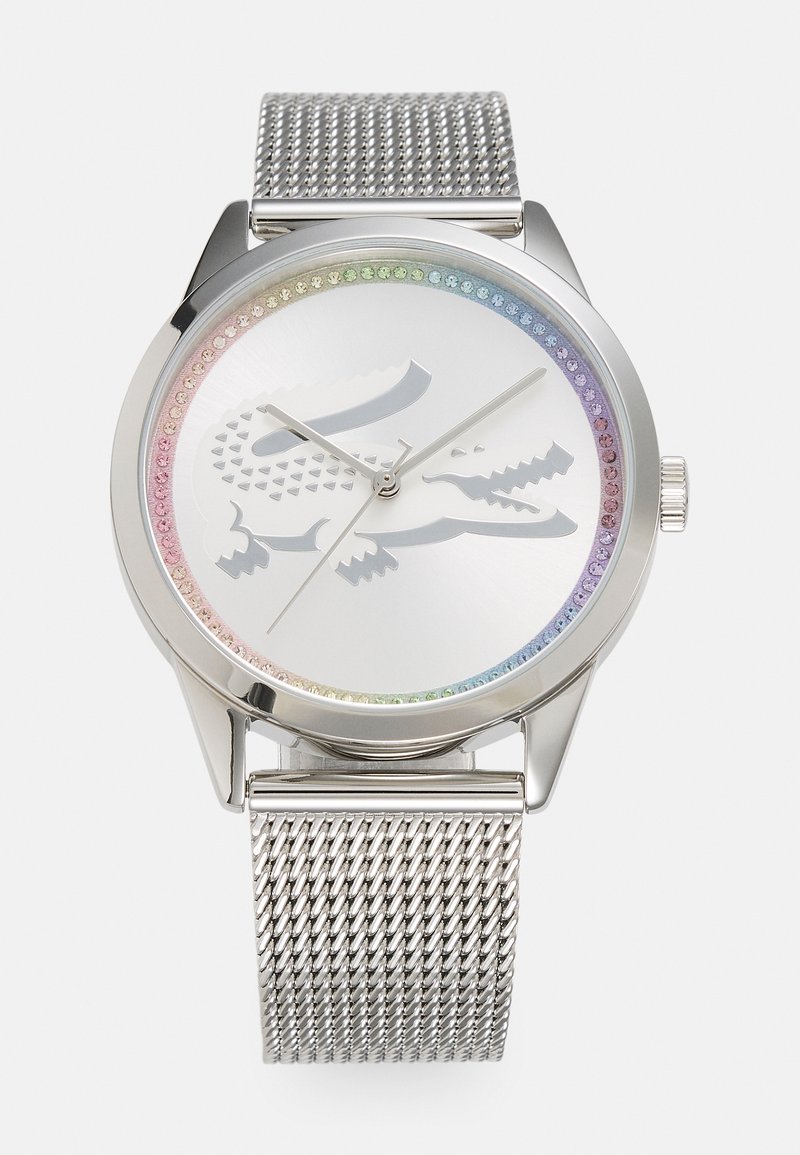 Reloj con correa de malla plateada y caja metálica. La esfera blanca cuenta con un bisel de cristal en tonos arcoíris y un sutil logo de cocodrilo en el centro.