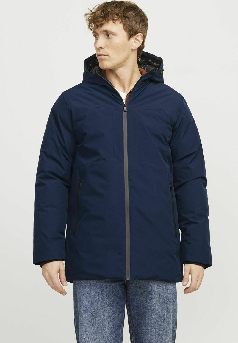 Veste isolante bleu marine avec fermeture éclair à l'avant, deux poches latérales et une capuche. Présente un design élégant et un tissu durable.