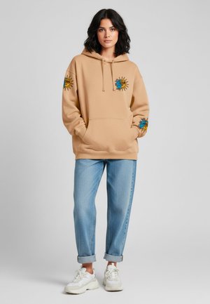 Felpa beige con cappuccio e ricami floreali a forma di sole sulle maniche, tasca a marsupio, abbinata a jeans azzurri arrotolati e scarpe da ginnastica bianche.