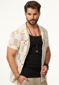 Kurzärmliges, beige gestricktes Shirt mit offener Struktur, getragen über einem schwarzen Tanktop. Auffällige braune Knopf-Akzente und übereinander getragene Halsketten.