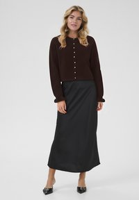 Rjava krajša cardigan jakna s podrobnostmi na gumbih, kombinirana z črno dolgo krilo z gladko teksturo in širšo zasnovo, dopolnjena s črnimi mule obutvami.