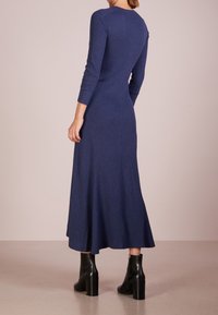 Robe bleu marine à manches longues en tissu texturé, design évasé atteignant le mollet. Portée avec des bottines noires à talons.