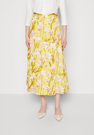 Femme portant une jupe midi jaune à imprimé floral rose, un haut blanc à manches longues et des escarpins crème à bout pointu avec des boucles dorées.