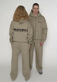 Beżowy komplet z kapturem i spodni dresowych z wyrazistym czarnym logo "MULTIPLY" z przodu i z tyłu. Miękka tekstura, luźny krój i ściągacz ze sznurkiem w pasie.