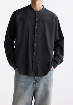 Hombre con camisa negra abotonada con cuello mao y vaqueros azul claro, de pie con las manos en los bolsillos contra un fondo blanco.
