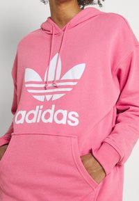 Rosa Adidas-hoodie med vit logotyp, kenguruficka, dragsko i huvan och ribbstickade muddarna. Mjuk textur och avslappnad passform.