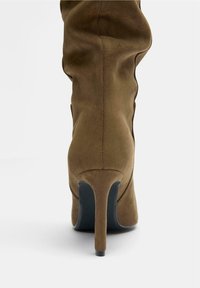 Bottine en suède marron avec un talon effilé, présentant une texture lisse et un design épousant la forme, mettant en valeur des contours élégants.