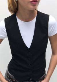 Veste noire ajustée avec un col en V profond, cinq boutons, et un tissu lisse, portée sur un t-shirt blanc. Caractérisée par un design élégant.