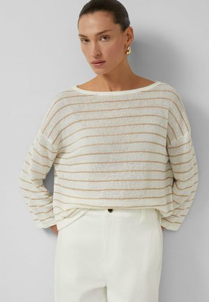 Kobieta z wygładzonym do tyłu włosami, nosząca złote kolczyki koła, biały sweter z cienkimi beżowymi poziomymi paskami i białe spodnie z wysokim stanem.