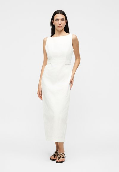 Γυναίκα με μακριά σκούρα μαλλιά που φοράει ένα λευκό αμάνικο midi φόρεμα και μαύρα σανδάλια με λουριά, στέκεται μπροστά σε ένα απλό φωτεινό φόντο.