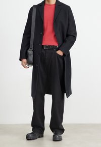 Lång svart ullkappa över en röd ribbad trikåtröja, i kombination med lösa svarta byxor och svarta klumpiga stövlar. Inkluderar en svart axelväska.