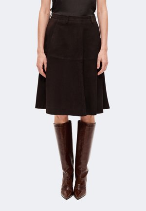 Femme portant une jupe noire longueur genou avec des poches et des bottes hautes marron foncé à bout pointu, debout devant un fond uni.
