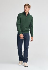 Pullover verde con zip a metà e piccolo logo, abbinato a jeans blu scuro e sneakers chiare. Tessuto liscio, vestibilità rilassata e maniche lunghe.