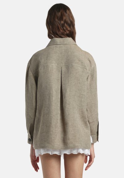 Femme aux longs cheveux bruns portant une veste ample en lin beige sur une jupe blanche à ourlet festonné, debout de dos devant un fond uni.