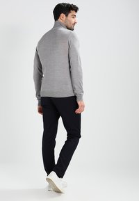 Grå strikket høyhalset genser med en tettsittende design, kombinert med mørke bukser og hvite sneakers, sett fra baksiden mot en enkel bakgrunn.
