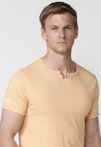 Svetle oranžové tričko s krátkým rukávem z měkké látky, kulatým výstřihem a dvěma knoflíkovými detaily, s malým logem na hrudi a na rukávu.