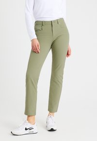 Person mit olivgrünen Slim-Fit-Hosen und weißen Nike Air Max Sneakers, der vor einem schlichten weißen Hintergrund steht.
