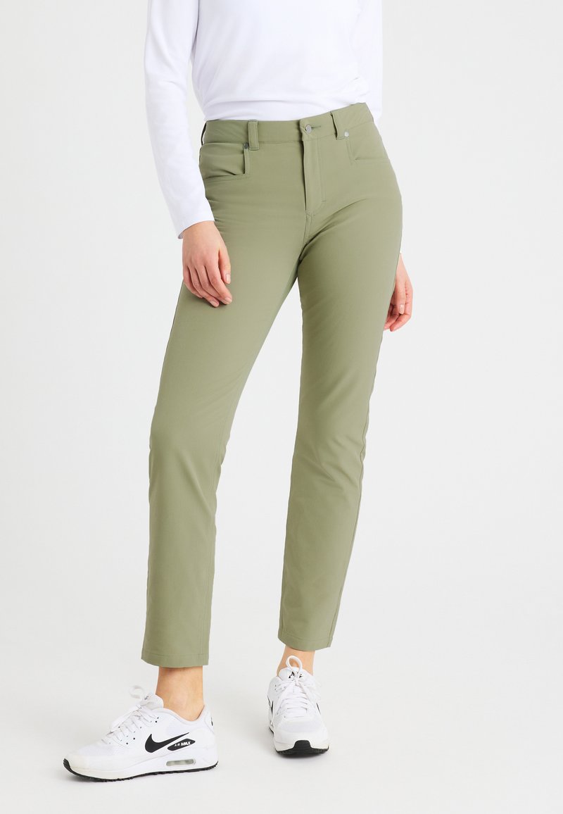 Person mit olivgrünen Slim-Fit-Hosen und weißen Nike Air Max Sneakers, der vor einem schlichten weißen Hintergrund steht.