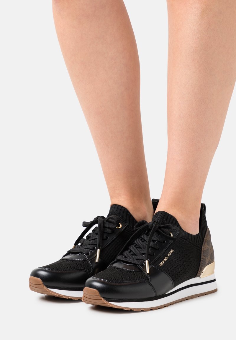 Black athletic shoes with a knit upper, leather accents, and a gold-tone heel detail. Features flat laces and a rubber sole.