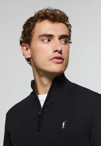 Svart zip-up tröja med ribbad krage, med en liten vit logga på bröstet. Tillverkad av ett mjukt stickat tyg.