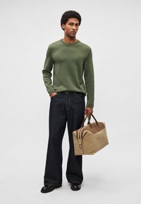 Camisola de malha verde com mangas compridas, jeans escuros e uma mala de pele bege com detalhes castanhos. Sapatos pretos completam o conjunto.