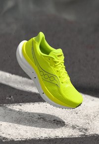 Kirkkaan keltainen juoksukenkä valkoisella pohjalla, ilmassa asvaltin yläpuolella, jossa näkyy Sauconyn logo ja kevyt verkkokuosi.