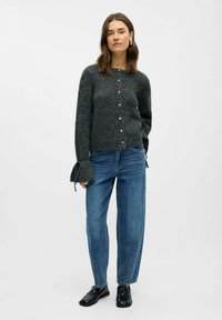 Grå strikket cardigan med perleknapper, flagrende ærmer, parret med lyseblå jeans og sorte loafers. Simpelt, afslappet stil.