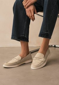 Beige slip-on shoes made of suede with a smooth finish and stitched accents. The sole is white, and the design is low-cut and minimalistic.