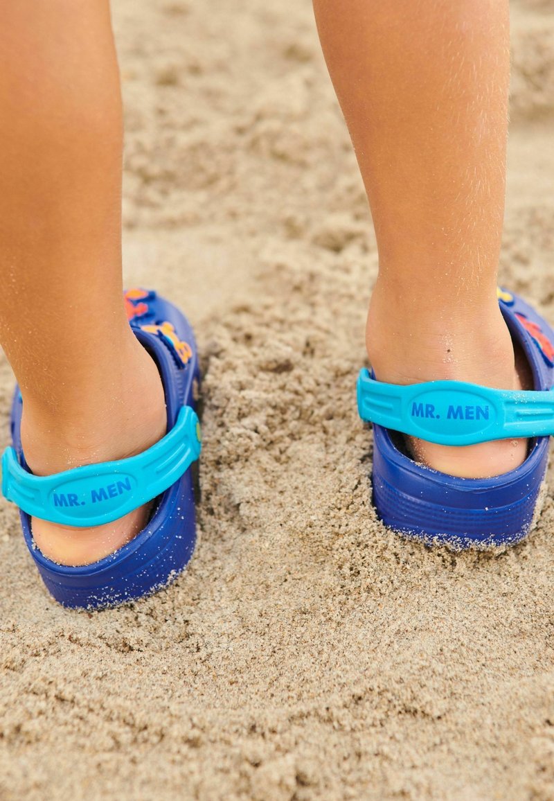 Sabots en plastique bleu avec une bride turquoise portant le texte « MR. MEN », partiellement enterrés dans un sable fin, présentant une texture lisse.