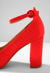 Chaussure à talon haut en suède rouge avec un talon bloc épais et une bride de cheville fixée par une boucle ronde, vue de côté.