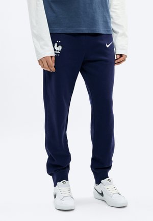 Persona vistiendo pantalones deportivos franceses azul marino con zapatillas Nike blancas y una camiseta de manga larga en una pose de pie.