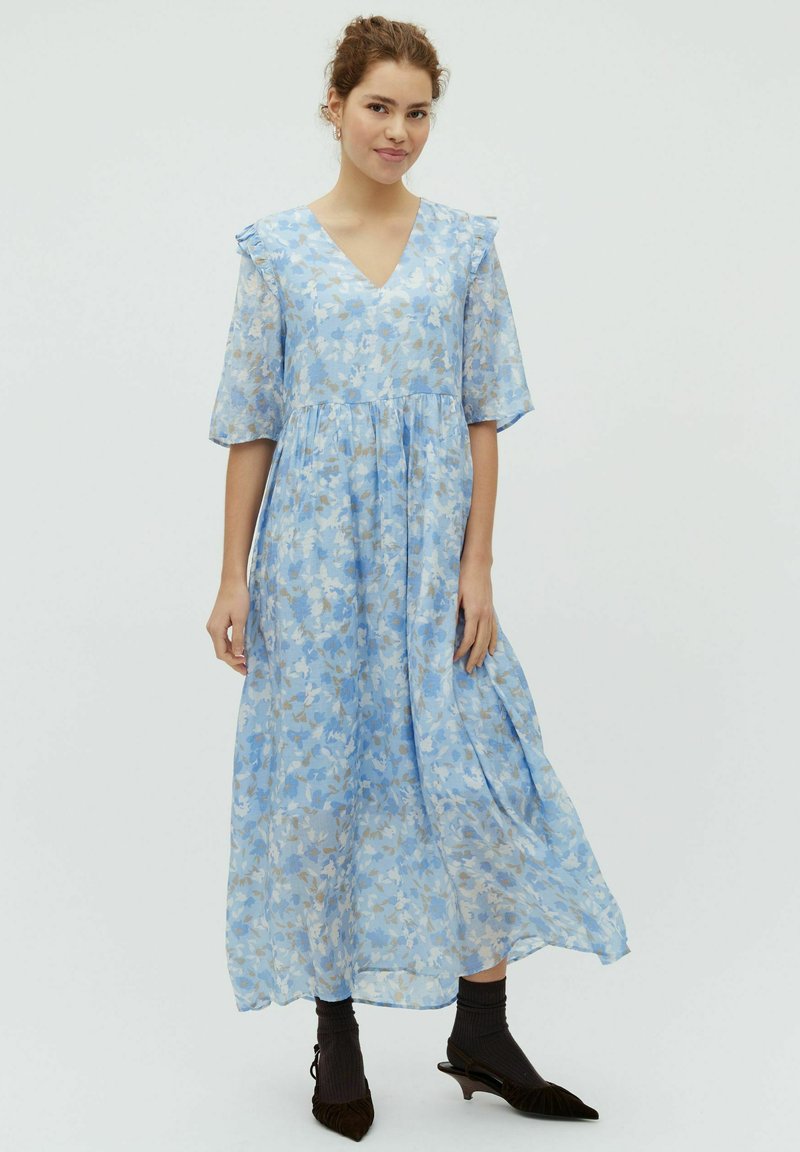 Jeune femme portant une robe midi fleurie bleu clair avec des épaules à volants, des chaussettes noires et des talons pointus noirs, debout devant un fond uni.