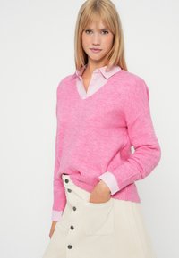 Kvinne med rett blondt hår, iført en rosa V-hals genser over en stripete rosa skjorte og en kremfarget knappe-skjørt, med hånden i lomma.