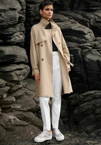 Beige trenchcoat met een dubbel knoopsgatdesign en een grote kraag, gecombineerd met witte op maat gemaakte broek en lichtgekleurde sneakers.