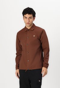 Giacca button-up marrone con colletto, polsini elastici e patch con logo. Realizzata in materiale liscio, presenta un taglio dritto e tasche laterali.