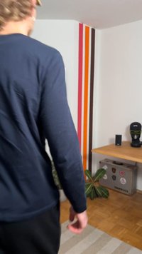 Person wearing a navy long-sleeve shirt and black pants, facing a room with a wooden table, potted plant, and colorful striped wall.