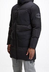 Doudoune noire avec capuche, dotée d'une fermeture éclair à l'avant, de deux grandes poches avant et d'un tissu texturé. Comprend un patch logo sur la manche.