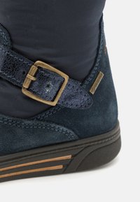 Sötétkék velúr bokacsizma, textúrált pánttal és bronz csattal, vízálló GORE-TEX címkével, és fekete gumi talppal, fából készült csíkokkal.