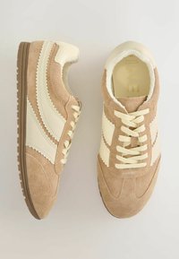 Bēšas zamša sporta kurpes ar krēmkrāsas sintētiskiem akcentiem, zigzaga detaļām un šņorzāļu aizdari. Teksturēta gumijas zole un mīksta iekšējā apdare.