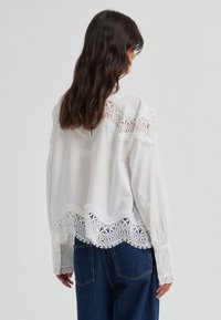 Blouse blanche avec des détails en dentelle sur le plastron et l'ourlet, manches avec poignets boutonnés et coupe décontractée. Portée avec un jean bleu foncé.
