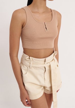 Frau trägt beiges, geripptes Croptop mit vorderen Hakenverschlüssen und hoch taillierte cremefarbene Paperbag-Shorts mit Gürtel, der in der Taille gebunden ist.