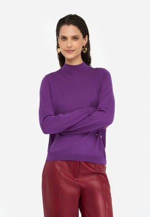 Maglione a collo alto viola, con maniche lunghe e una texture morbida, dotato di leggere aperture laterali, abbinato a pantaloni in finta pelle rossa profonda.