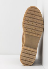 Semelle d'une chaussure marron clair avec des rainures diagonales et de petites indentations circulaires, appuyée contre un mur et une surface blancs.