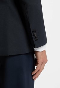 Giacca di abito blu navy con finitura testurizzata, caratterizzata da una vestibilità slim, quattro bottoni neri sul polsino e un polsino di camicia bianca che spunta sotto.