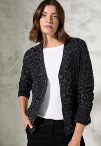 Černý žebrovaný cardigan s bílými skvrnami, má kulatý výstřih, dlouhé rukávy a zapínání na pět knoflíků. Nosený přes bílou košili.