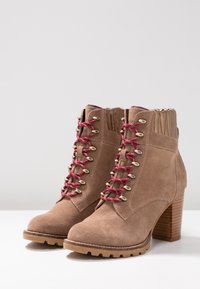 Brązowe botki zamszowe z czerwono-białymi sznurowadłami w wzory, złotymi przelotkami i teksturowanym drewnianym obcasem. Wytrzymała podeszwa zapewniająca przyczepność.